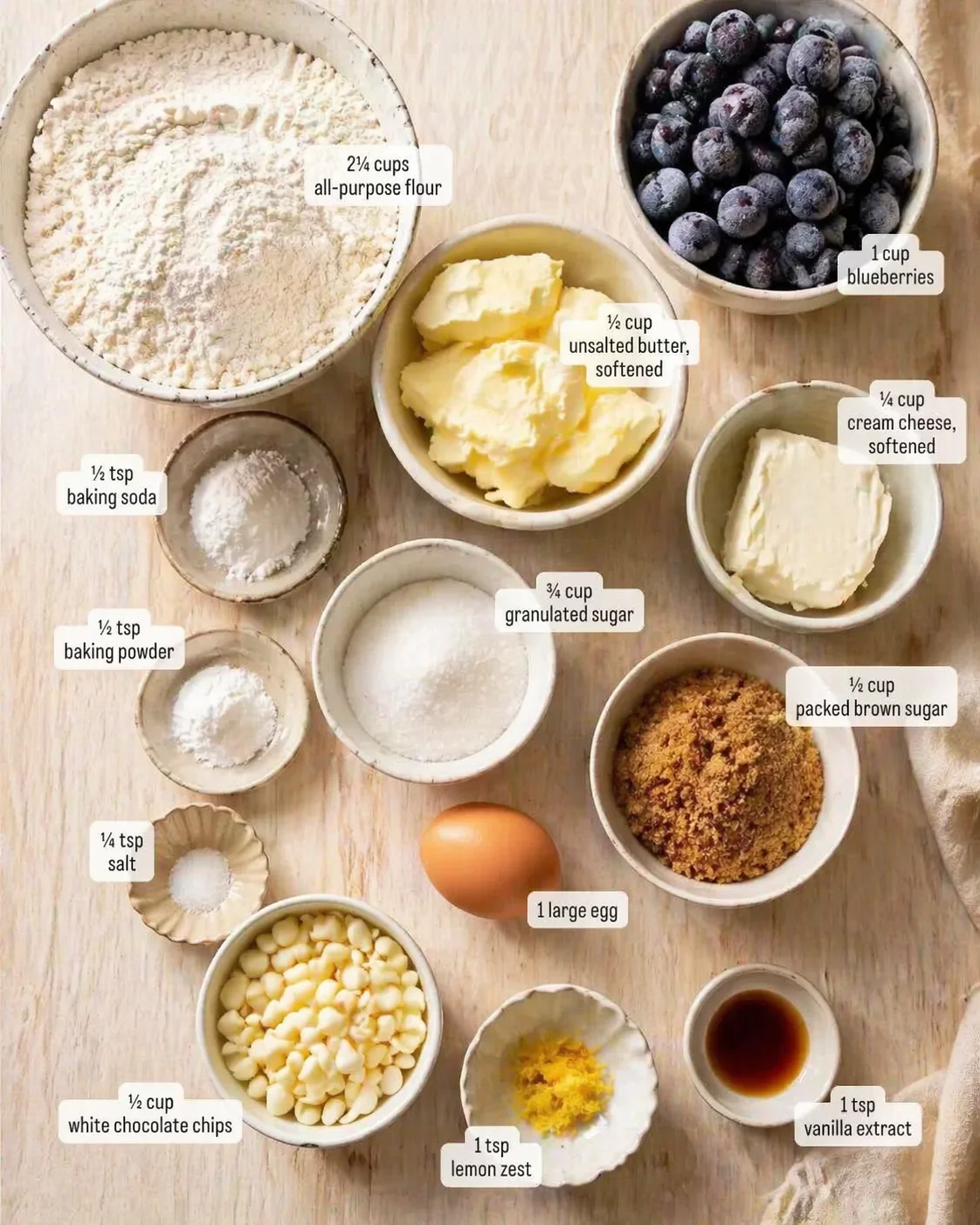 Overhead view of a baking ingredients layout with flour, butter, blueberries, sugars, and eggs on a wooden table.
