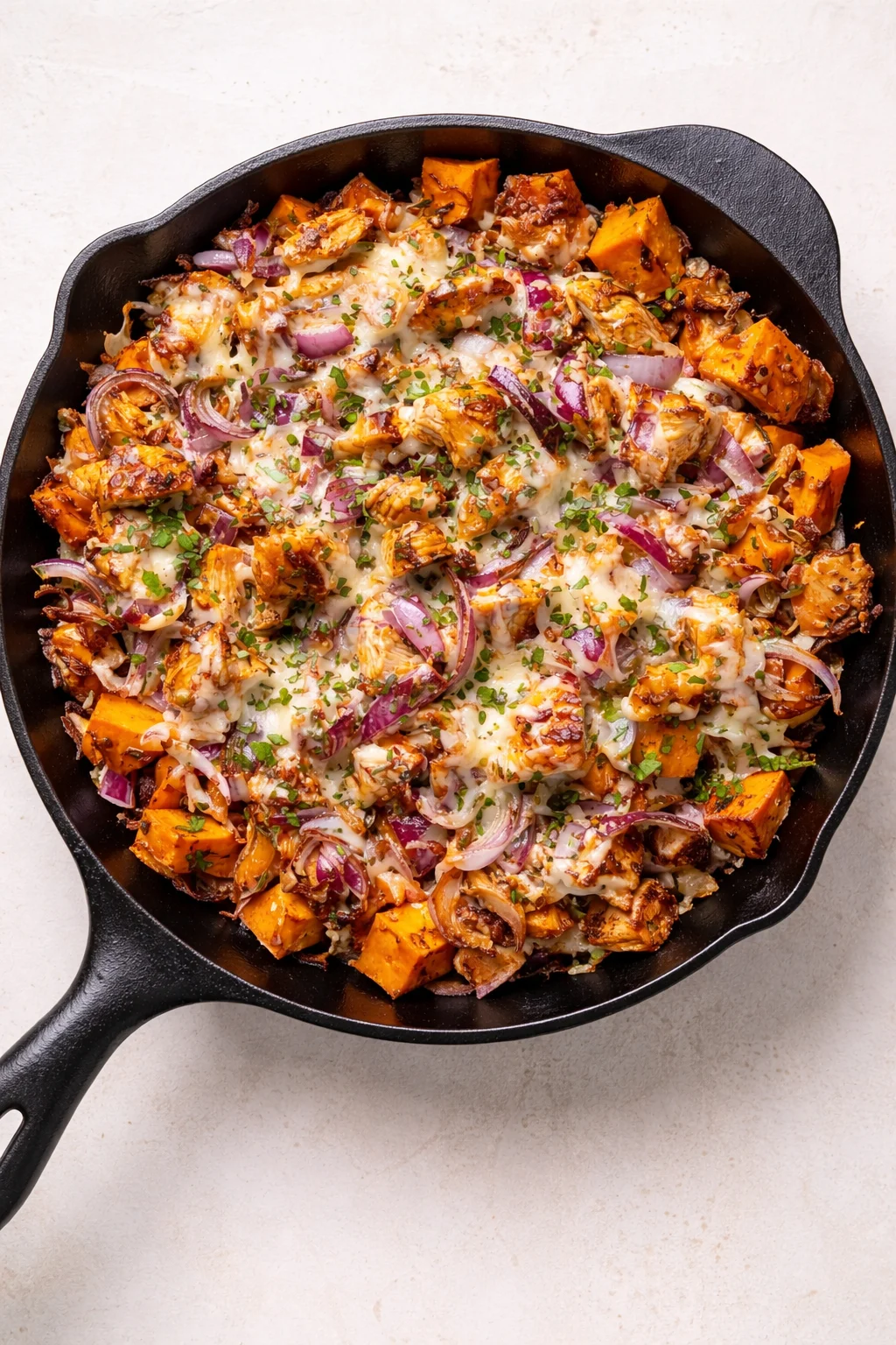 A cast-iron skillet filled with roasted sweet potato cubes, red onions, melted cheese, and herbs.