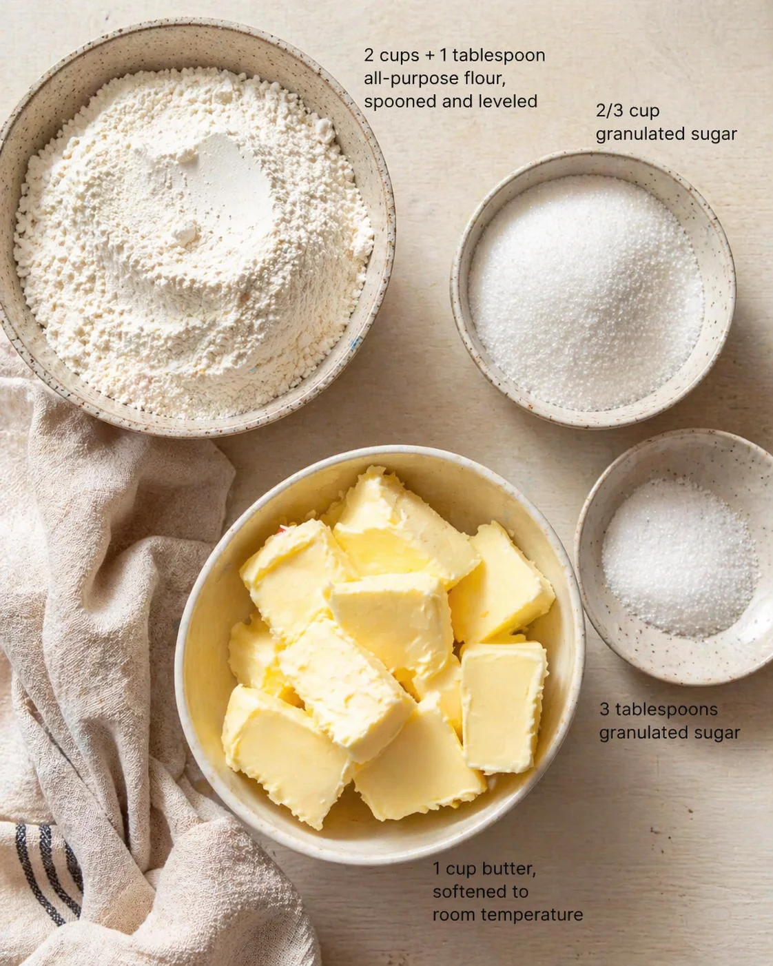 Top-down view of bowls containing flour, sugar, and butter with measurement labels on a beige wooden surface.