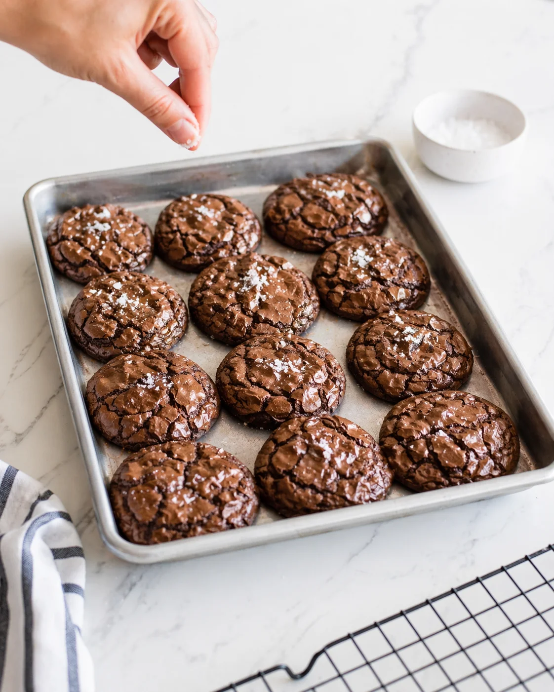 Tray of chocolate crinkle cookies on a marble counter, salt being sprinkled on top.