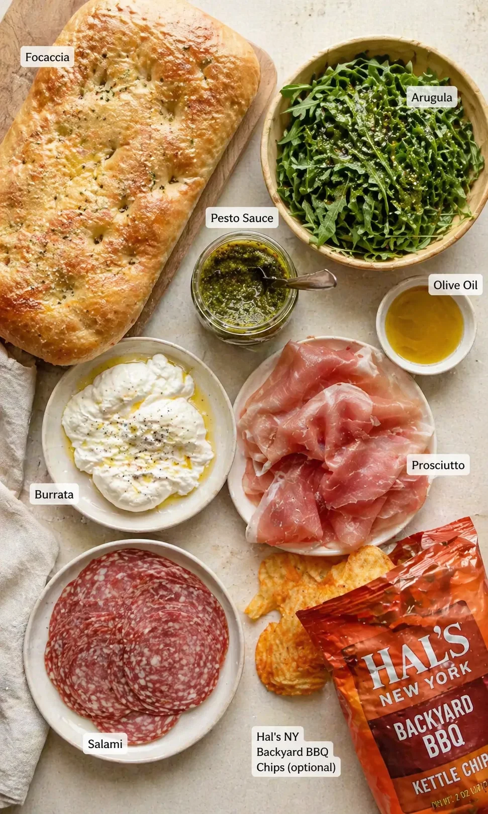 top-down view of a prep board with focaccia, burrata, arugula, prosciutto, salami, pesto, olive oil, and chips