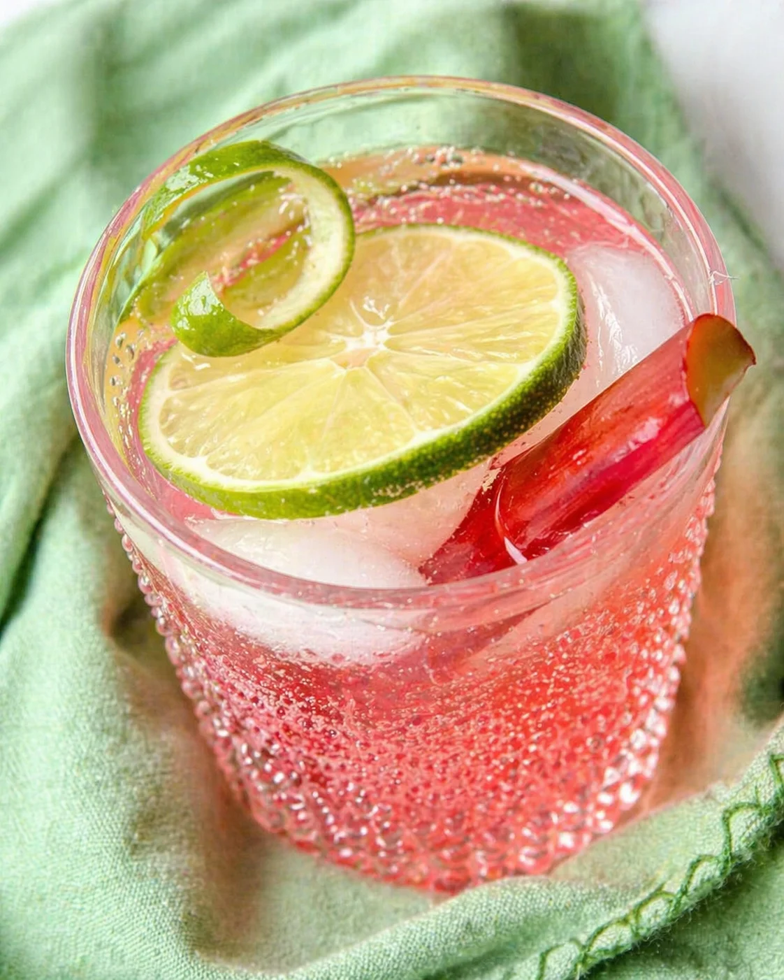 A pink citrus drink in a beaded glass with lime wheel garnish on a green linen cloth