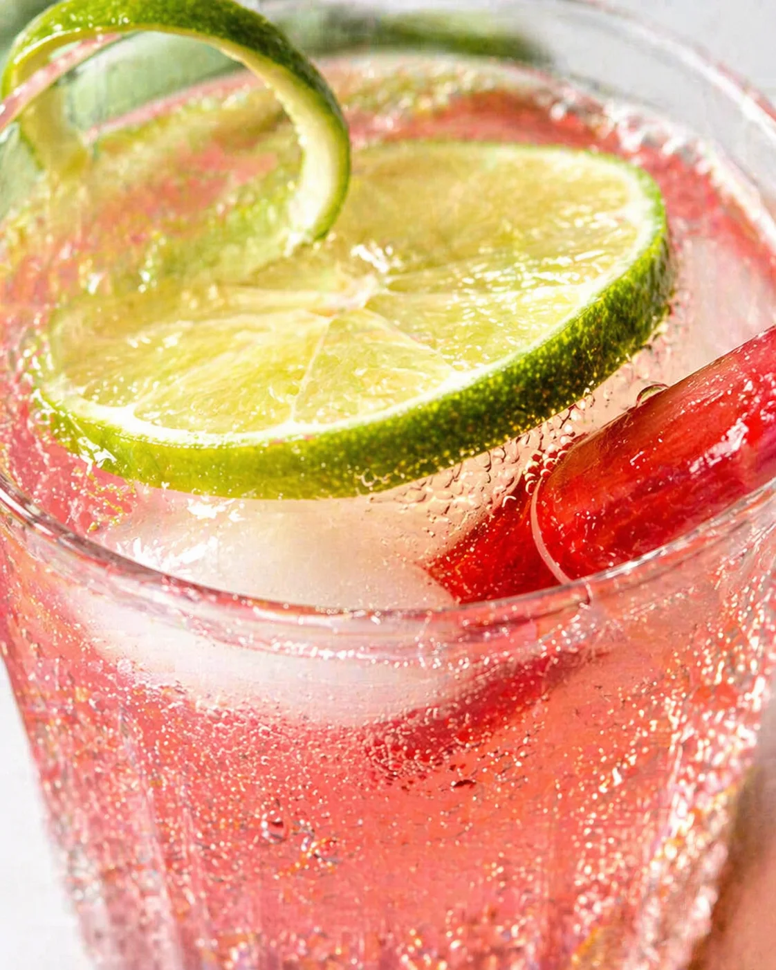 Close-up of a pink sparkling drink in a glass with lime wedge and cucumber garnish