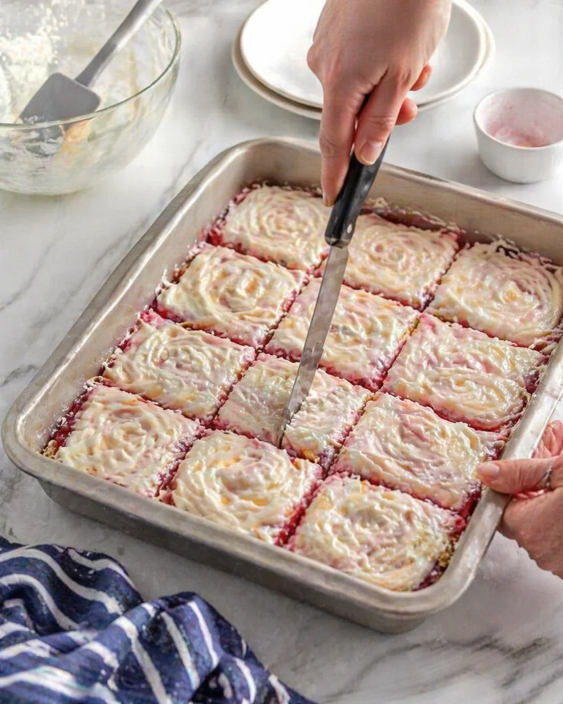 hand slicing pink frosted cheesecake bars in a metal baking pan on marble countertop