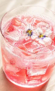 Close-up of a pink drink in a glass with ice and white flower garnish.