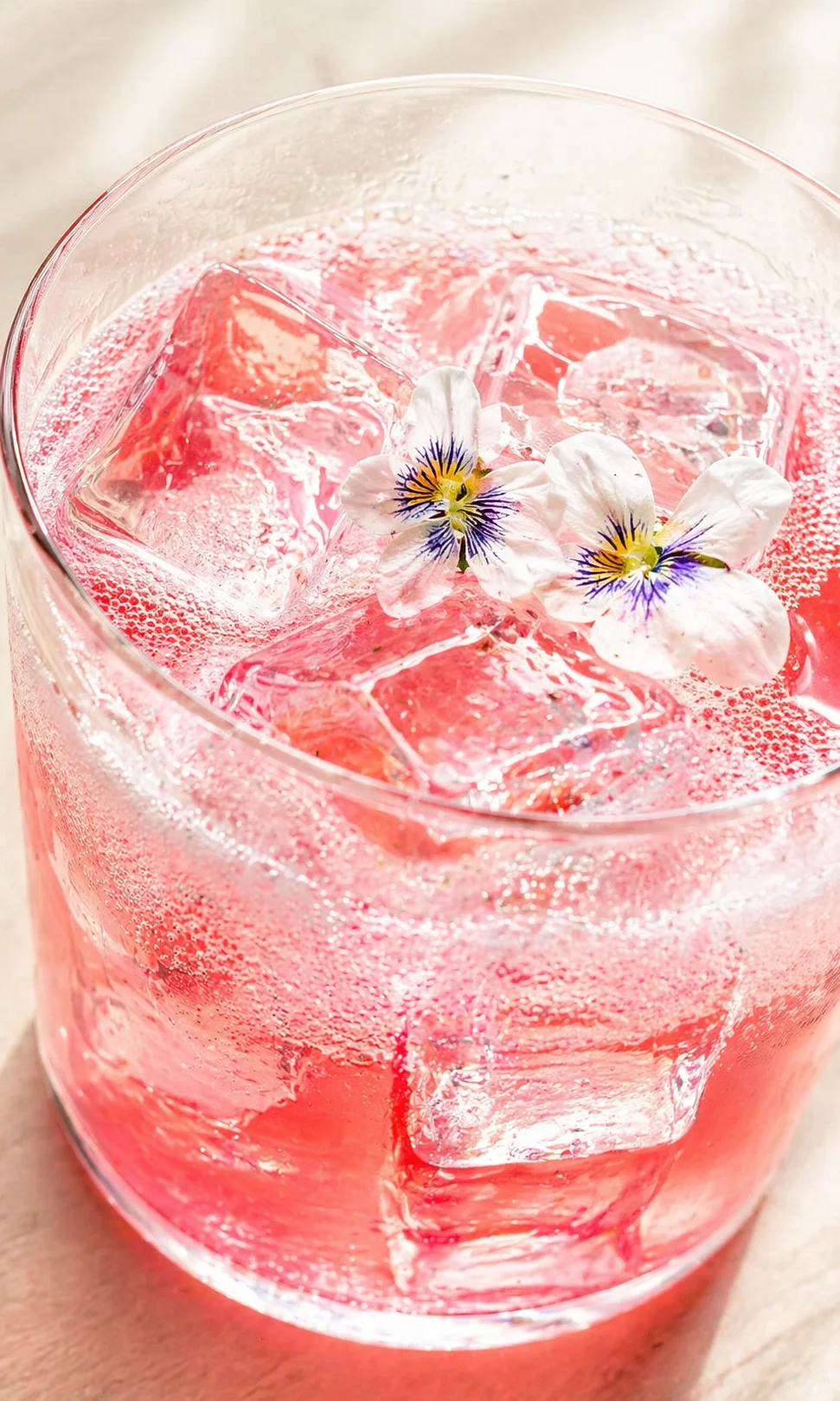 Close-up of a pink drink in a glass with ice and white flower garnish.
