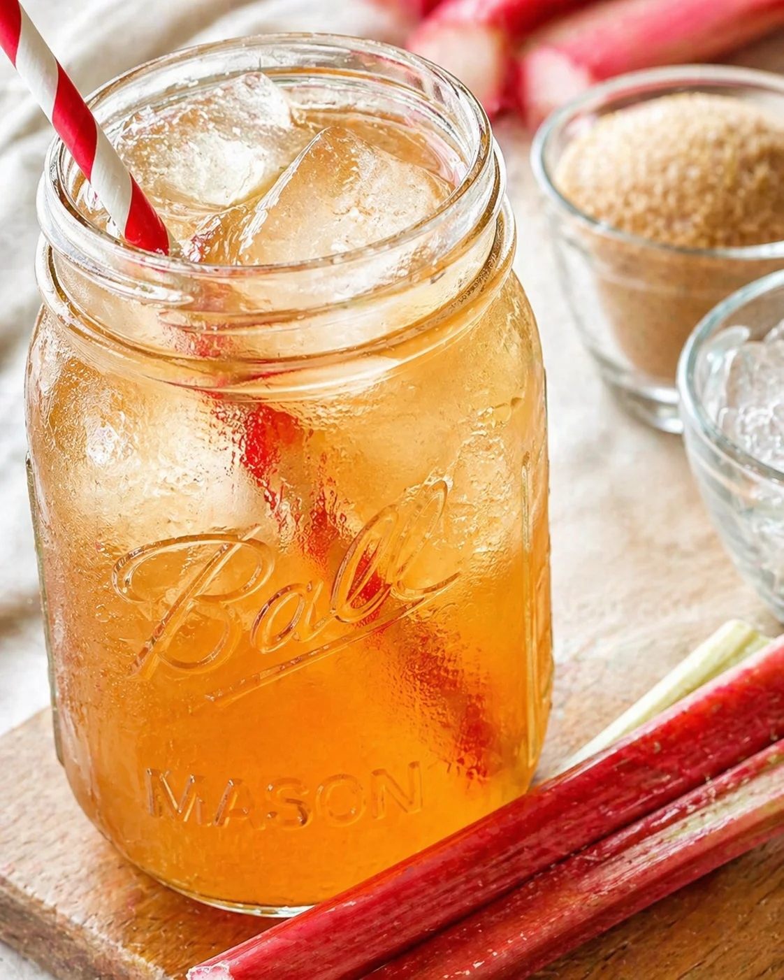 Amber iced tea in a mason jar with a red-and-white striped straw, surrounded by rhubarb stalks.
