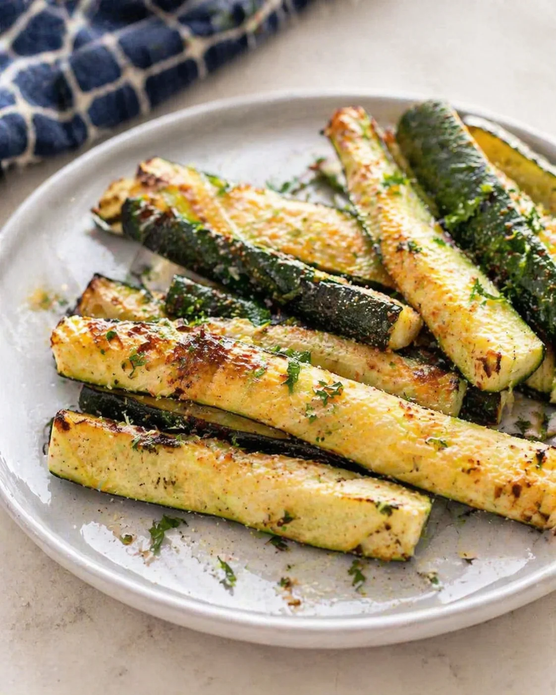 Plate of crispy roasted zucchini fries sprinkled with parmesan and herbs on a light plate.