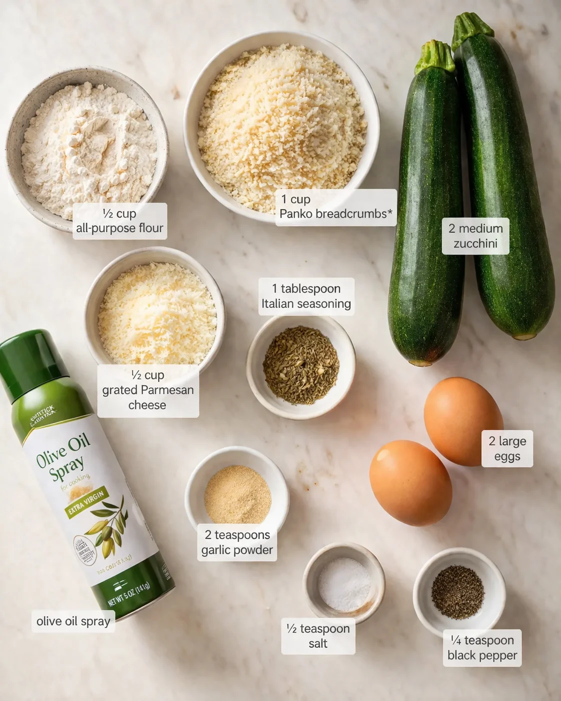 Overhead view of ingredients laid out on marble: zucchinis, eggs, flour, breadcrumbs, cheese, spices, olive oil spray.