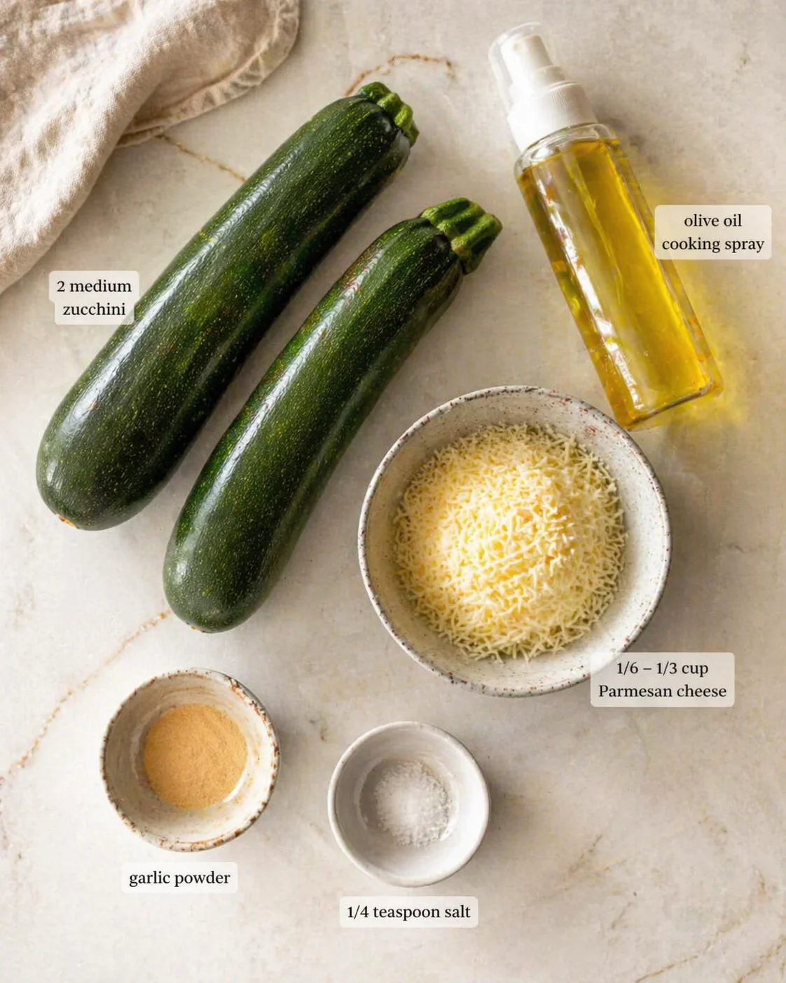 two dark-green zucchinis next to grated parmesan, olive oil spray, garlic powder, and salt on a light marble surface