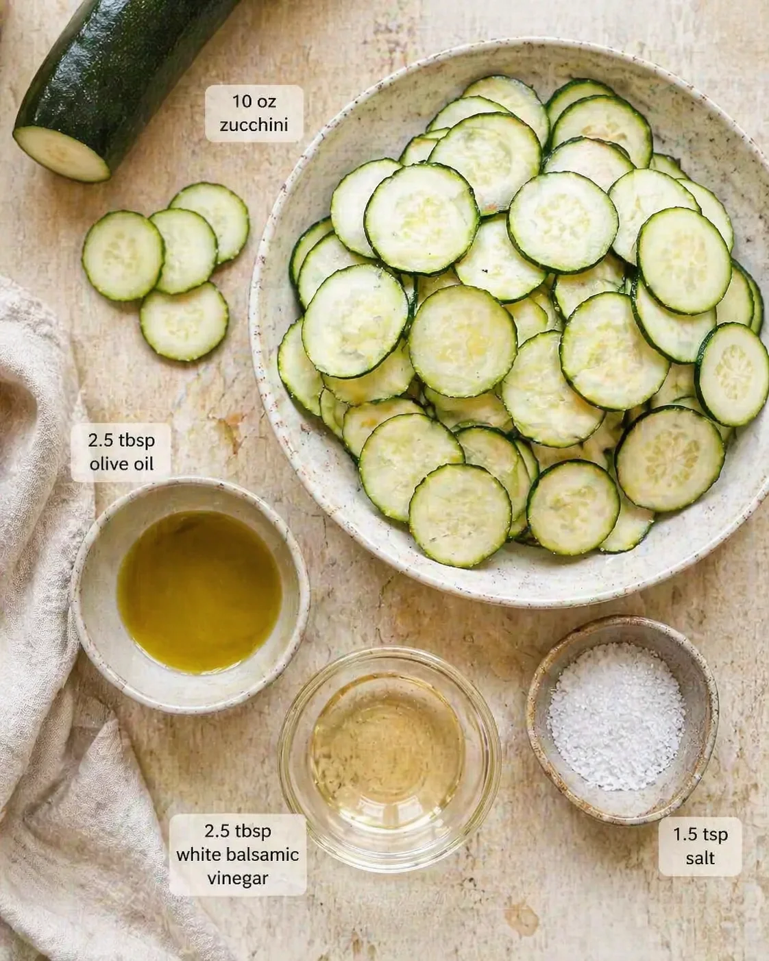 Top-down shot of thinly sliced zucchini arranged in a bowl with olive oil, white balsamic vinegar, and salt nearby.