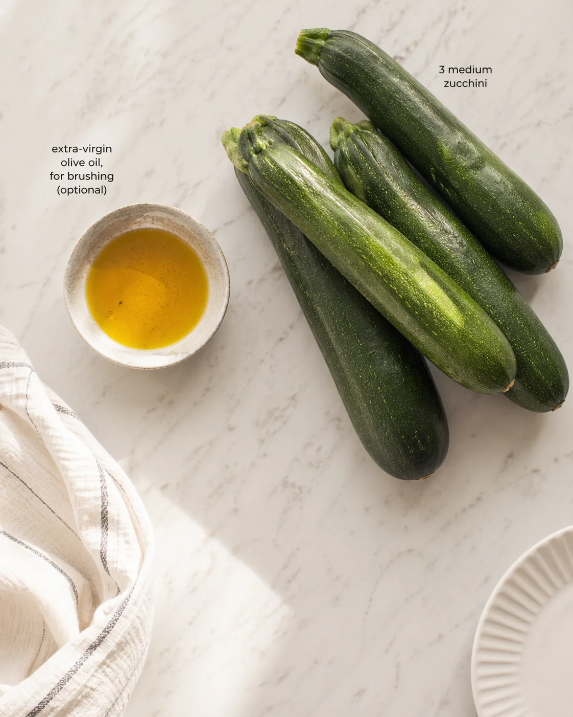 Three medium zucchinis and a bowl of olive oil on a marble countertop with recipe labels.