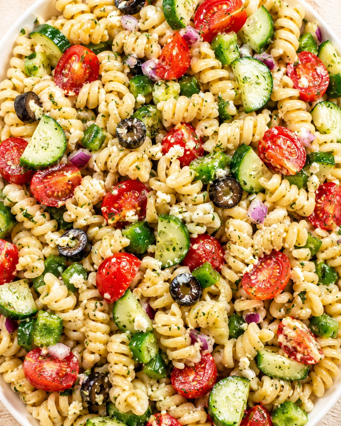 Close-up of creamy fusilli pasta salad with tomatoes, cucumber, olives, red onion, and feta.
