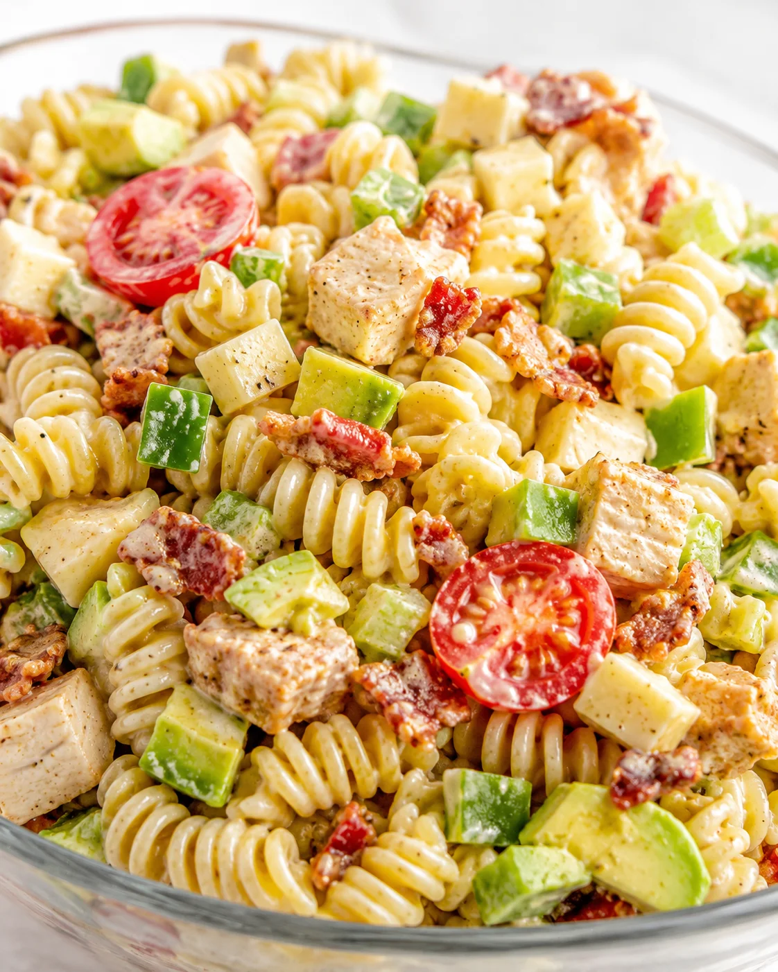 Creamy fusilli pasta tossed with chicken, avocado, bacon bits, and cherry tomatoes in a glass bowl.
