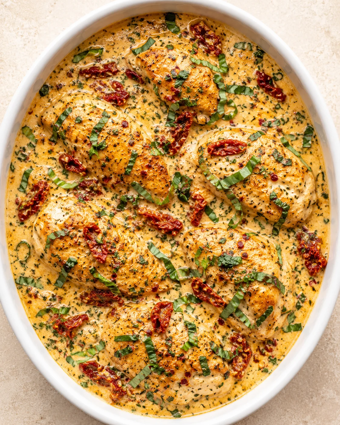 oval white dish with chicken in creamy sauce, sun-dried tomatoes, and chopped herbs