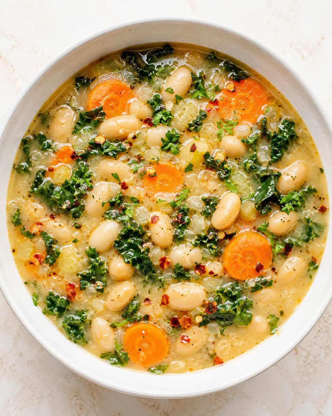 a creamy white bean soup with carrot rounds, kale, and herbs in a white ceramic bowl.