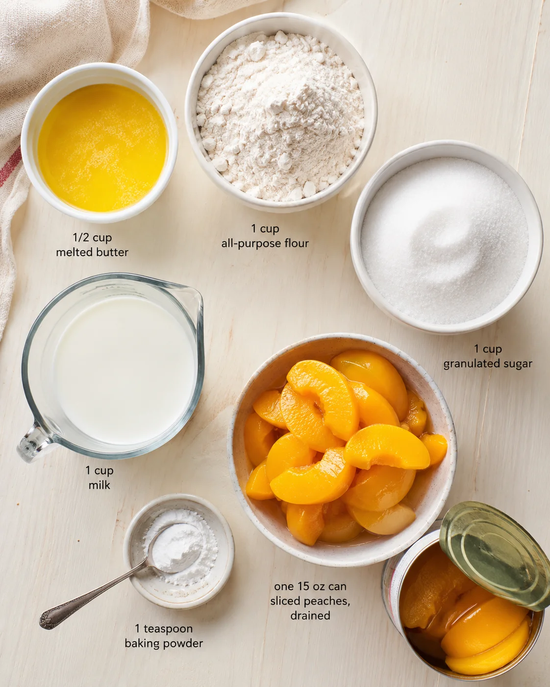 Top-down view of peaches and baking ingredients: melted butter, flour, sugar, milk, baking powder, on a pale wooden surface.
