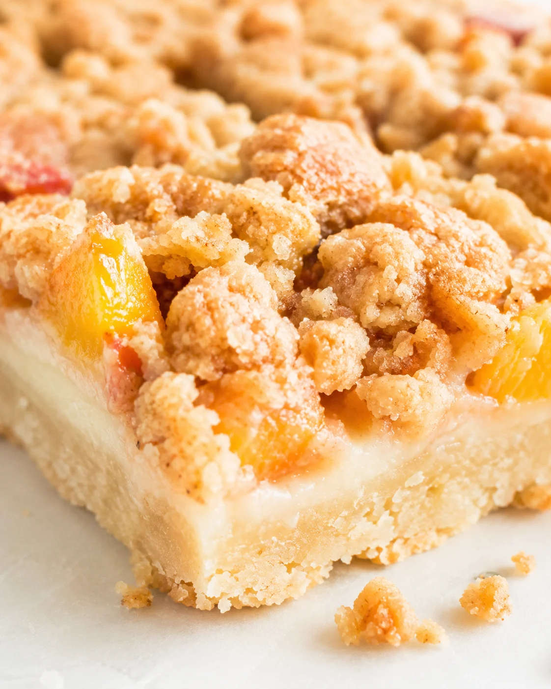 Close-up of a peach crumble bar with crumbly topping and peach chunks on a light surface.