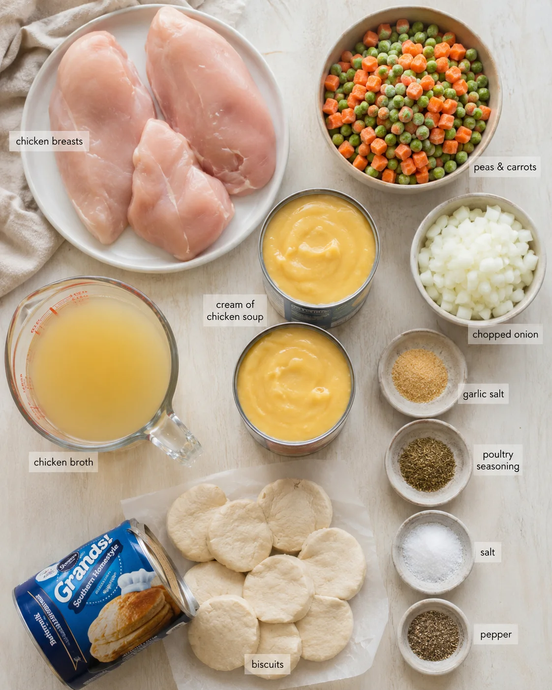 Overhead view of chicken breasts, vegetables, canned soup, broth, biscuits and seasonings arranged on a light wooden surface.