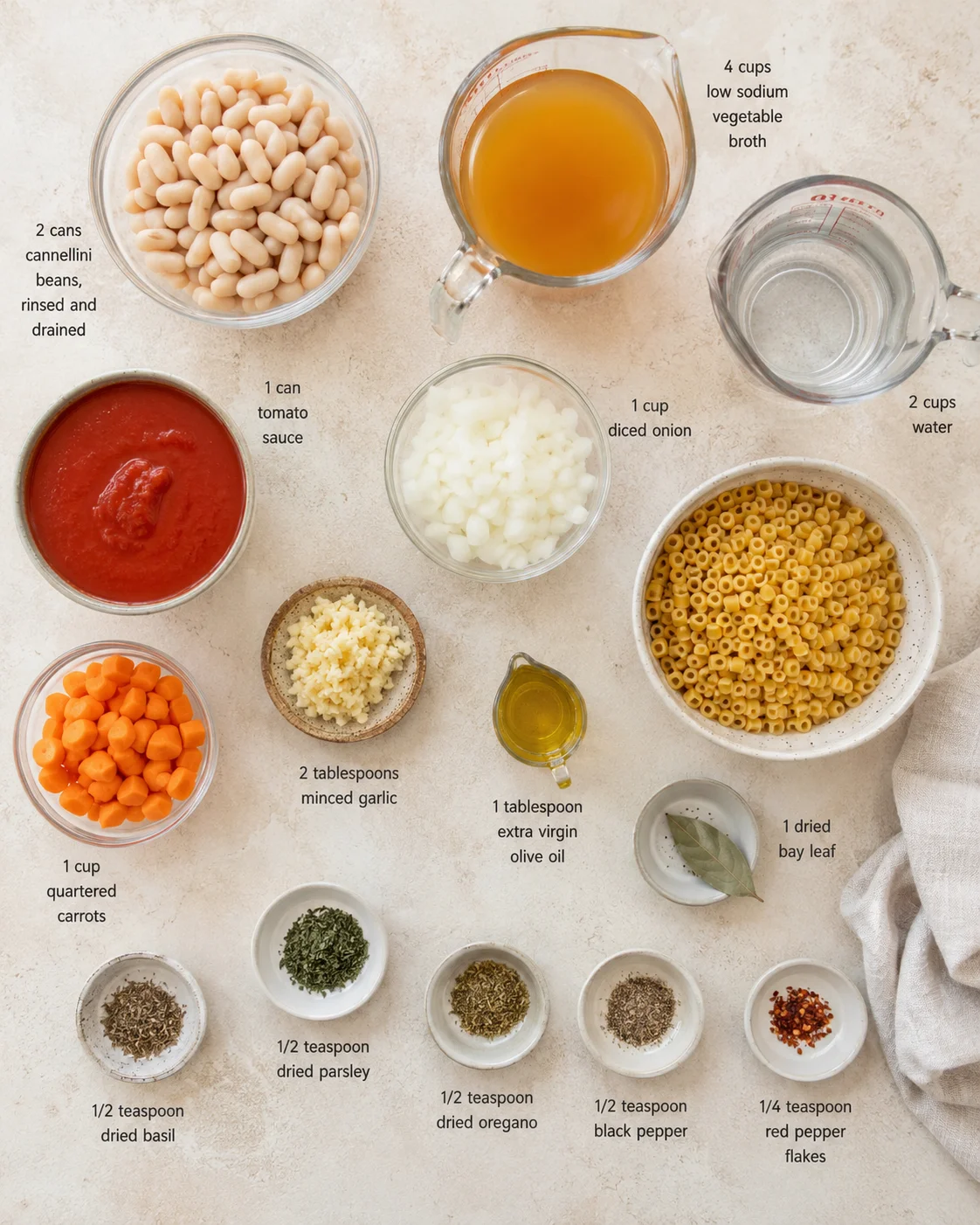 Overhead flatlay of organized pasta ingredients arranged on light countertop with beans, sauce, broth, vegetables, and herbs.