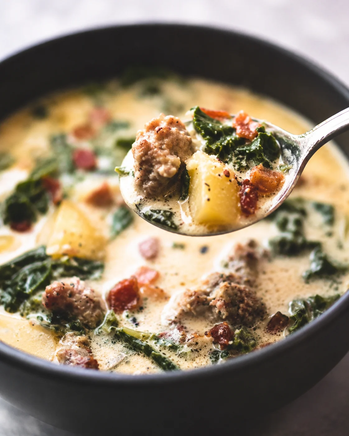 Spoon lifting a spoonful of Olive Garden Zuppa Toscana showing sausage, potato, and kale above a bowl